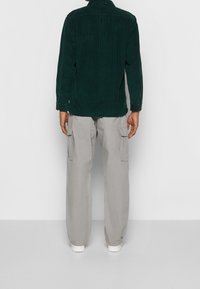 Camicia in corduroy verde con colletto, maniche lunghe e finiture testurizzate, abbinata a pantaloni cargo grigi chiari con tasche laterali.
