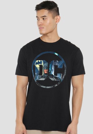 Junger Mann, der ein schwarzes T-Shirt mit blauem Batman-Logo und den Buchstaben DC vorne trägt und nach links schaut.