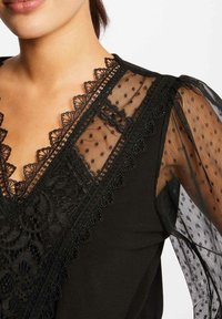 Blouse noire avec des manches ballon transparentes à pois. Présente un délicat bord en dentelle autour du col en V et des accents en dentelle décoratifs sur les manches.