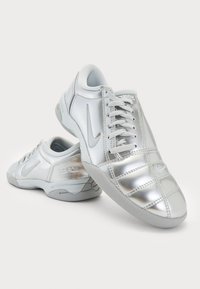 Zapatillas sintéticas plateadas con textura suave, diseño de cordones y cuello acolchado. Presenta un logotipo y una suela de goma gris en contraste.