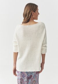 Hvid løsstrikket sweater med bred halsudskæring og rullekanter, parret med en skørtepants i paisley-mønster. Stoffet ser blødt og chunky ud.