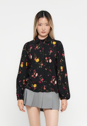 Modella che indossa un blouse lungo a fiori nera con maniche a sbuffo e una gonna mini plissettata grigia, su uno sfondo bianco semplice.