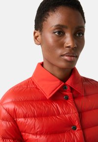 Rote Steppjacke mit Kragen, die horizontale Steppmuster und schwarze Knopfakzente aufweist. Glatte, glänzende Stofftextur.