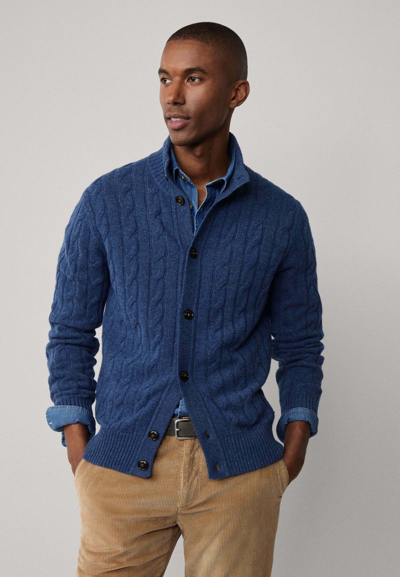 Marineblauwe kabelgebreide cardigan met een sjawl kraag, voorzien van zwarte knopen en geribde manchetten. Draagt over een lichtblauw shirt en tan corduroy broek.