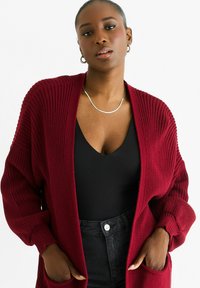 Burgundy knitted cardigan with a loose fit, open front, and ribbed texture. Worn over a black top and paired with high-waisted black jeans.