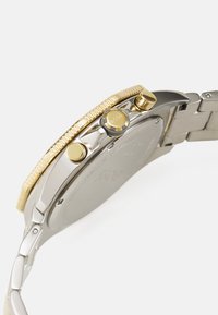 Reloj de pulsera en tonos plateado y dorado que presenta un bisel texturizado, tres botones de presión y una correa de acero inoxidable con un acabado pulido.