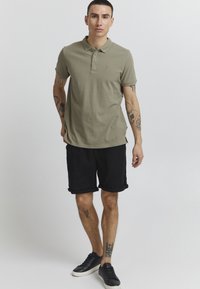Oliven grønn polo skjorte i bomull, med krage og tre knapper, kombinert med svarte shorts. Brukt med svarte sneakers, har tatoveringer på armene.