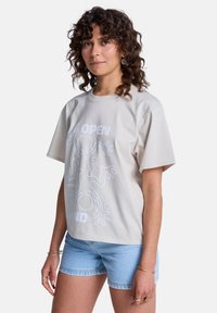 Μπεζ βαμβακερό μπλουζάκι με σχέδιο σε λευκή γραμμή που απεικονίζει χέρια και αφηρημένα σχήματα, σε συνδυασμό με ανοιχτόχρωμα μπλε σορτς από τζιν.