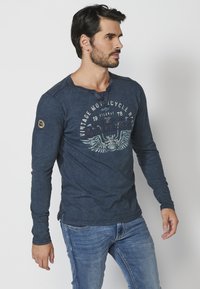 Dlouhý rukáv, tmavě modré tričko s výstřihem henley, grafický potisk zobrazující křídla a text, a logo nášivka na levém rukávu.