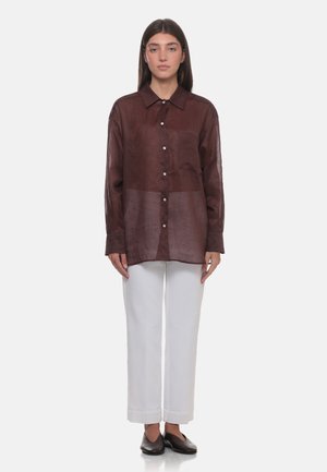 Donna in piedi che indossa una camicia marrone trasparente a maniche lunghe con bottoni, pantaloni bianchi a gamba larga e scarpe basse marrone scuro su sfondo bianco.