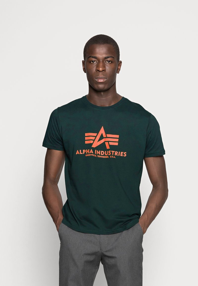 Mørkegrøn T-shirt med et orange trykt logo og teksten "ALPHA INDUSTRIES," rund halsudskæring, korte ærmer og et tætsiddende design.