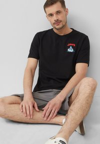 Schwarzes Baumwoll-T-Shirt mit "JAWS" Grafik auf der Brust, kombiniert mit grauen Shorts. Modell sitzt, Hände ruhen auf den Beinen, neutraler Hintergrund.