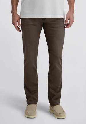 Man met slim-fit bruine broek, beige instapschoenen en een wit shirt, staand tegen een eenvoudige lichte achtergrond.