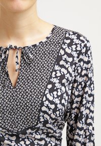 Blusa floral navy con escote tipo ojal y lazo, mangas estampadas y suaves impresiones florales en azul claro que contrastan. Tela suave y ligera.