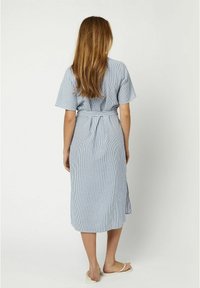 Donna con capelli lunghi che indossa un vestito midi a righe blu e bianche con cintura e infradito bianchi, in piedi contro uno sfondo neutro con la schiena rivolta verso l'osservatore.