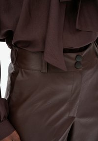 Zbliżenie na ciemnobrązowe skórzane spodnie z wysokim stanem, z czarnymi guzikami oraz dopasowaną brązową bluzką z wiązaniem w pasie.