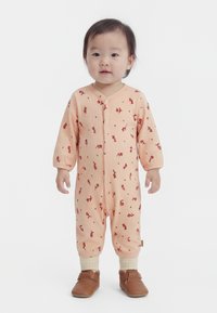 Małe dziecko stojące w brzoskwiniowym body w kwiaty, kremowych skarpetkach i brązowych miękkich bucikach na białym tle.