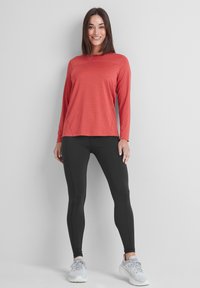 Woman standing and smiling, wearing a red long-sleeve top, black leggings, and white running shoes against a plain light background.