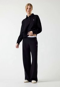 Zwarte trui met een kraag en drie knopen, gecombineerd met losse zwarte broek. Beide zijn gemaakt van een zachte stof. Witte sneakeraccenten zichtbaar.