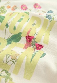 Texte vert-jaune indiquant « BORN » sur un tissu floral avec des feuilles vertes et des fleurs roses brodées sur un fond beige.