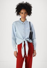 Ljusblå denimskjorta med en knut i midjan, knäppning fram, långärmad och broderad logotyp. Känns perfekt ihop med röda byxor.