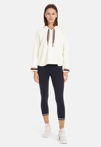 White hooded sweatshirt with a loose fit and ribbed texture, paired with navy leggings featuring a brown and white stripe at the ankle.