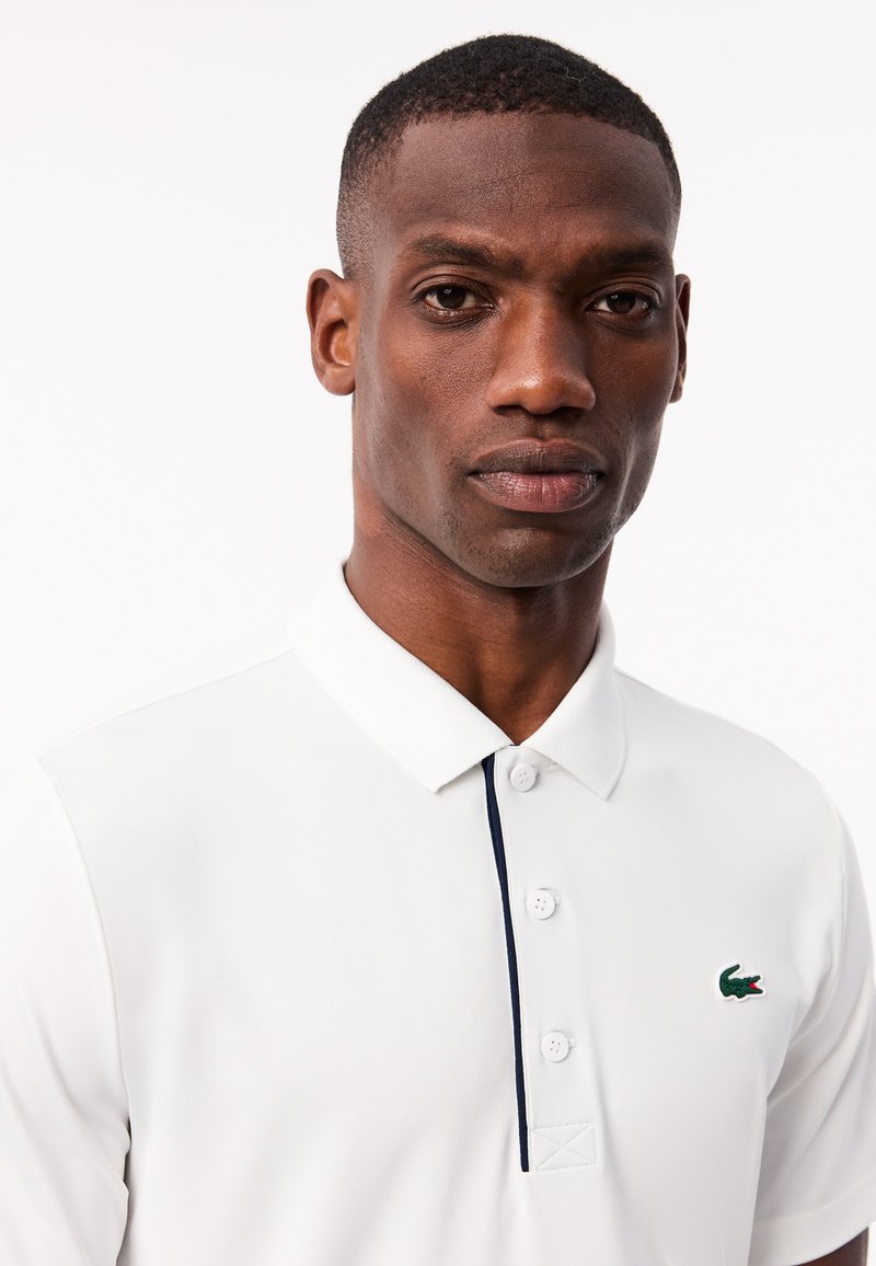 Polo shirt blanc en mélange de coton, avec une patte de boutonnage à trois boutons, accent marine sur le col et logo brodé vert sur la poitrine.