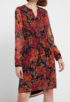 Kvinde iført en knælange sort kjole med røde, orange og blå blomsterprint, knaplukning foran, lange gennemsigtige ærmer og bånd i taljen.