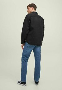 Camisa negra de algodón, con cuello y puños de botón, combinada con jeans azules desgastados y zapatillas negras. Fondo liso.