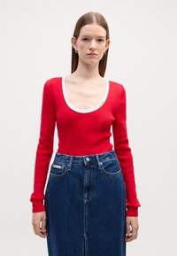 Rød langermet topp med hvit scoop-hals, kombinert med en mørk denimskjørt med frontslit og merkeinformasjon på linningen.