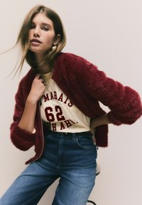 Jachetă din blană sintetică burgundă, tricou crem cu imprimat "MARAIS 62", blugi din denim albastru cu talie înaltă și colier minimalist din aur.