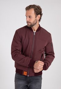 Giacca bomber bordeaux con polsini e orlo a costine, chiusura frontale con zip e tasca laterale con zip. Presenta un'etichetta arancione sull'orlo.