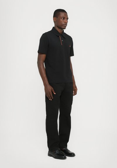 Polo noir avec col, manches courtes et un petit logo rouge sur la poitrine, associé à un pantalon noir et des chaussures habillées noires.