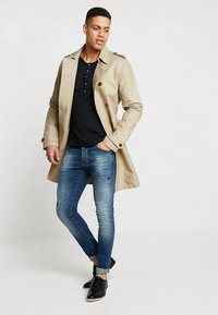 Trinchado beige con detalles en los botones, camiseta henley negra, jeans de mezclilla desgastados y zapatos de cuero negros. Pose de pie.