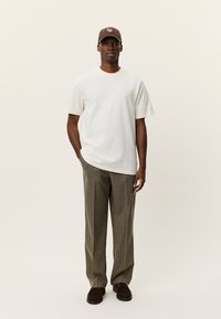 Μπλούζα από βαμβάκι σε ανοιχτό μπεζ με κοντά μανίκια, συνδυασμένη με καρό παντελόνι σε καφέ αποχρώσεις και καφέ καπέλο με λογότυπο. Παπούτσια σε βαθύ καφέ χρώμα.