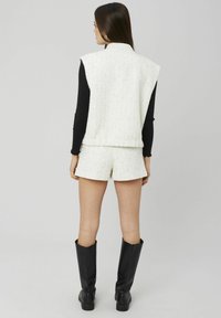 Veste blanche texturée sans manches associée à un short assorti. Chemise noire à manches longues en dessous et bottes noires montantes jusqu'aux genoux portées.