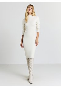 Donna con capelli biondi lunghi che indossa un abito a collo alto color crema aderente e stivali abbinati al ginocchio, in piedi davanti a uno sfondo chiaro e semplice.