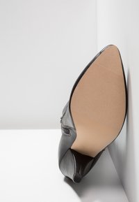 Schwarze Lederknöchelstiefel mit spitzer Zehe und niedriger Absatz, die sohlenseitig gegen eine weiße, eckige Fläche abgestellt sind.