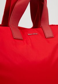 Bolso de lona rojo de nailon con asas de cuero. Presenta el logotipo en relieve "Marc O'Polo". Textura suave, forma rectangular y colores de asas contrastantes.