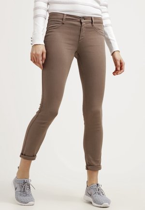 Person iført opsmøgede brune skinny bukser, hvid langærmet top med striber og grå Nike-sneakers, stående på en hvid baggrund.