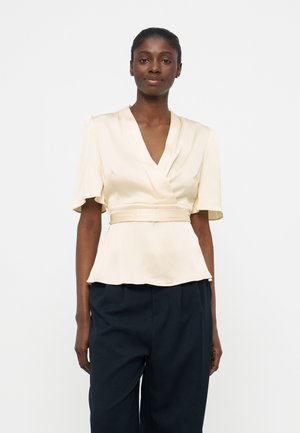 Γυναίκα που φοράει μπλούζα κρεπ σε κρεμ απόχρωση με κοντά μανίκια που φουντώνουν και εφαρμοστή μέση, συνδυασμένη με φαρδιά μαύρη παντελόνα, πάνω σε λευκό φόντο.