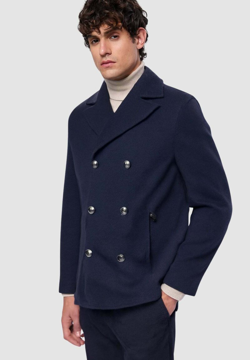Cappotto doppiopetto in lana blu navy con bottoni argentati, ampi revers e tasca laterale, abbinato a un dolcevita chiaro.