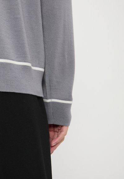 Pullover grigio con un design in maglia testurizzata, caratterizzato da una striscia bianca all'orlo. Il polsino è leggermente largo, poggiando su un indumento nero.