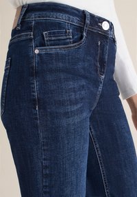 Gros plan sur une personne portant un jean en denim bleu foncé avec des coutures et des poches visibles, associé à un haut à manches longues blanc.