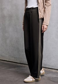 Pantaloni larghi e scuri con vita elastica, caratterizzati da strisce laterali beige a contrasto. Abbinati a sneakers bianche e marroni.
