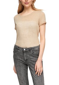 Beige kortärmad topp i lätt, texturerad tyg; har rundad halsringning och avslappnad passform, kombinerad med mörkgrå jeans.