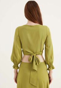 Blusa verde oliva con maniche lunghe a sbuffo, polsini arricciati e dettaglio con lacci sul retro, abbinata a una gonna coordinata. Tessuto morbido e leggero.