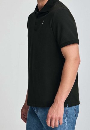 Homme portant un polo à manches courtes noir avec col et un jean bleu, debout devant un fond clair uni.