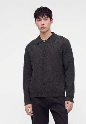 Jeune homme portant un pull gris foncé à boutons avec col et un pantalon noir, debout devant un fond clair uni.