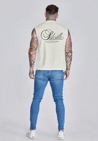 Top sin mangas de algodón color crema con texto negro del logo en la parte posterior, combinado con jeans ajustados azules y zapatillas blancas. Brazos tatuados visibles.